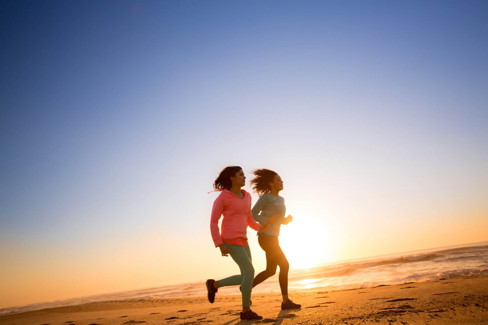 Two women practicing self-care by running on the beach at sunset for improved mental health.