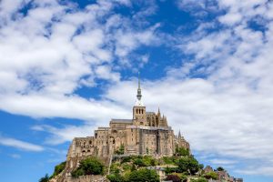 A historic stone abbey is perched atop Mont Saint-Michel, surrounded by lush greenery and under a partly cloudy blue sky. This iconic location often draws visitors who marvel at its beauty, much like the marathon runners who pass through its scenic trails every year.