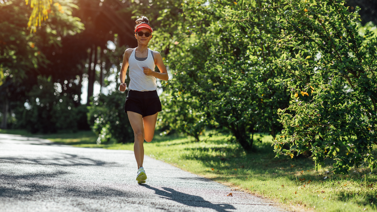 A woman in sportswear and a red cap jogging on a sunny park pathway, surrounded by lush greenery, engaging in her summer training.