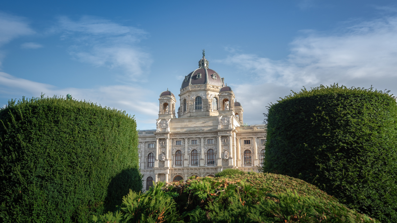 A historic building with a central dome stands under a blue sky, framed by neatly trimmed bushes in the foreground, reminiscent of Vienna's architectural beauty.