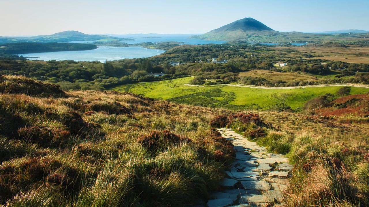 A stone pathway leads through grassy hills toward a distant mountain, with a lake and scattered houses in the background under a clear blue sky, reminiscent of the scenic route many take during the Connemarathon race.