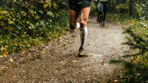 Two runners are depicted on a muddy trail in a forest, with one runner visible from the waist down wearing shorts and white compression socks, likely training for their first ultramarathon.