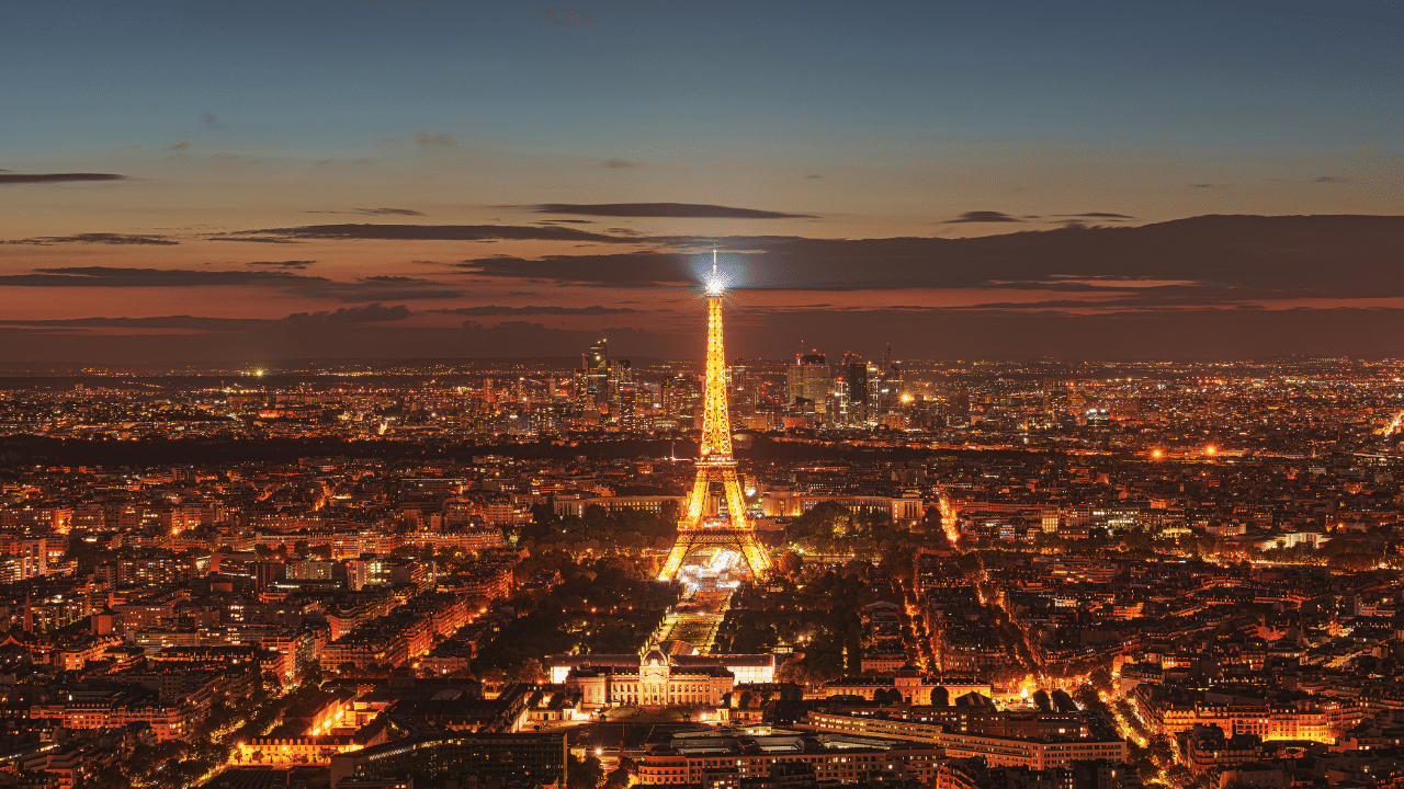 Aerial view of the Eiffel Tower in Paris illuminated at night, surrounded by buildings and city lights with a darkening sky in the background, capturing the spirit of Marathon Pour Tous as it winds through the city's iconic landmarks.