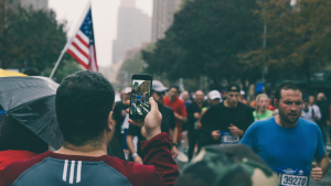 A person in a red jacket films a marathon using a smartphone. Runners, including one with bib number 39270, pass by as an American flag is seen in the background amidst the cityscape of this popular running race.