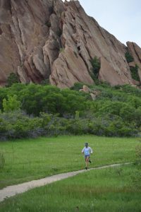 A person in a blue hat and white shirt runs along a dirt path through a green, grassy field with large rock formations and trees in the background, as if participating in one of the many running races held throughout the year.